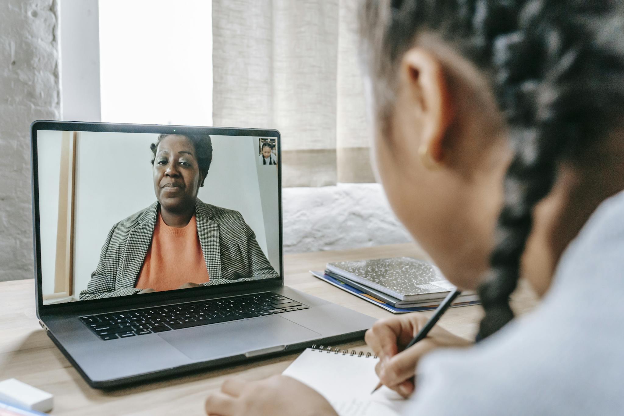 A girl participating in an online learning session with a tutor via video call on her laptop, taking notes.