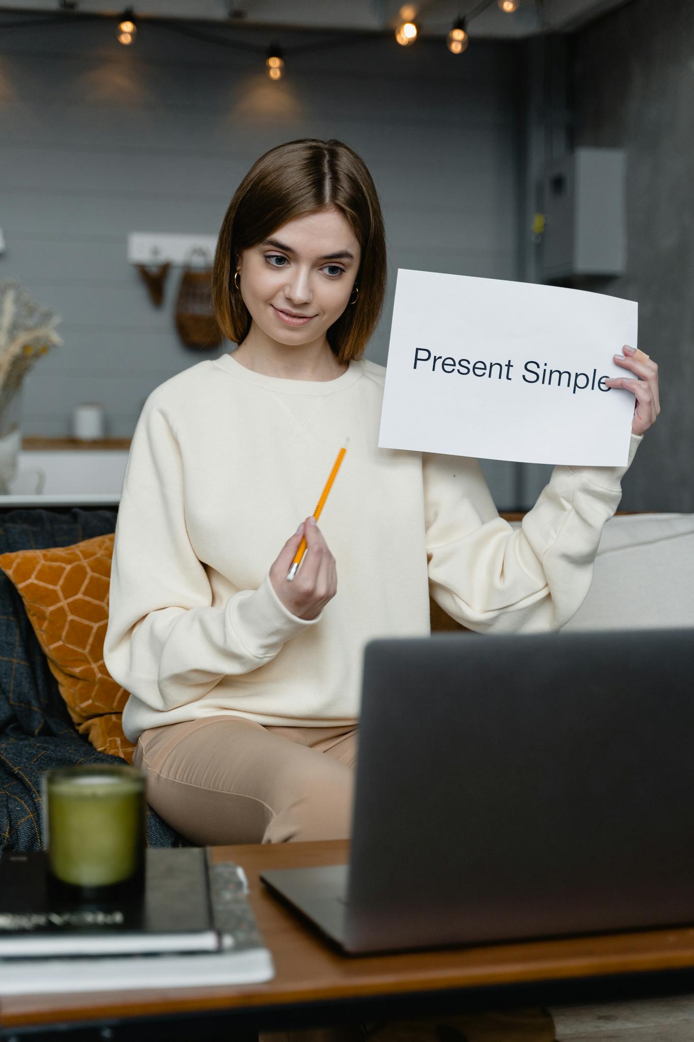 Woman teaching English online holding 'Present Simple' flashcard, engaging in video call.